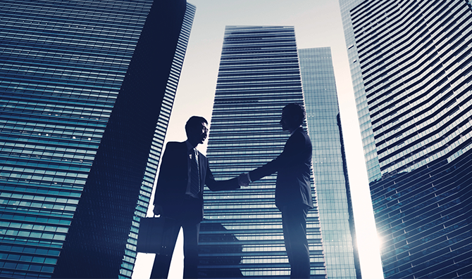 都会の高層ビルを背にビジネスマン２人が握手を交わしている写真